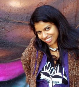 Image of a person standing against a wall, smiling. They have dark hair and are wearing a necklace, an animal print jacket, purple hoodie, and black t-shirt.