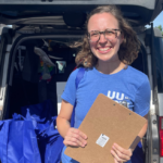 Nora Rasman, a woman with shoulder-length brown hair, smiling warmly and rocking a blue UU the Vote t-shirt. She is holding a clipboard and standing in front of an open car trunk in a parking lot. The sky is a clear blue and palm trees are visible in the background.