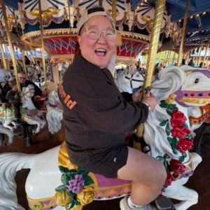 Josie Mitz is smiling energetically and riding a colorful horse on a carousel.